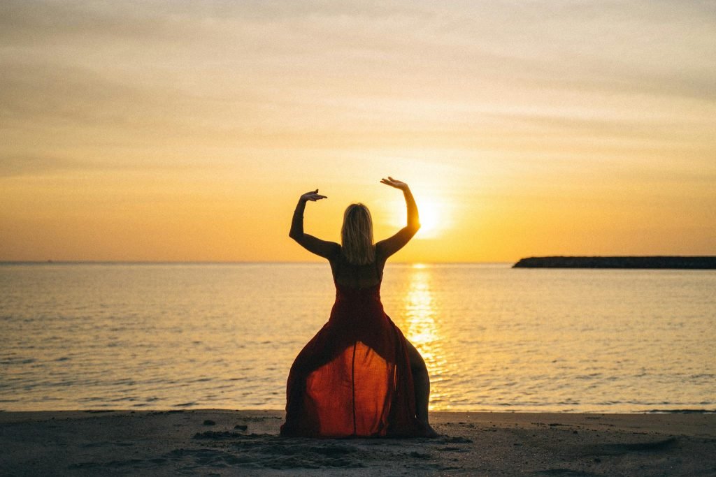 Back view of female with raised hands doing yoga standing on sandy beach with face towards calm sea and sunset in summertime
