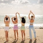 Five women in colorful activewear practicing yoga on a sunlit beach, raising arms joyfully.