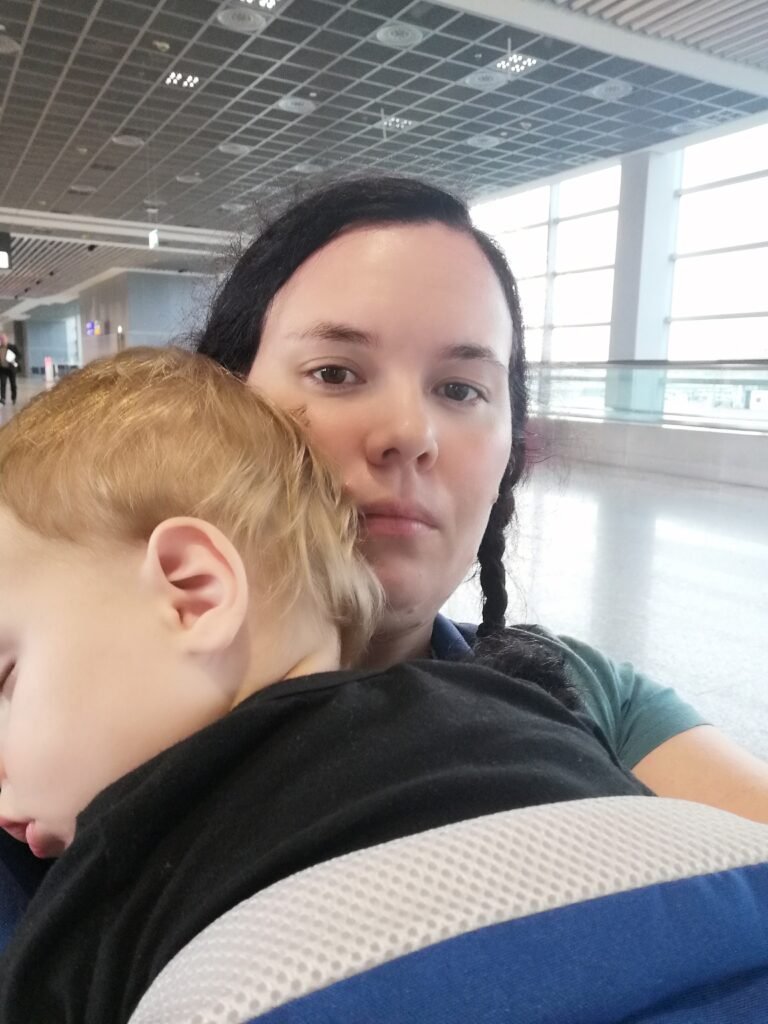 A selfie of a women with a plait holding a sleeping toddler in an airport during a layover in Amsterdam.