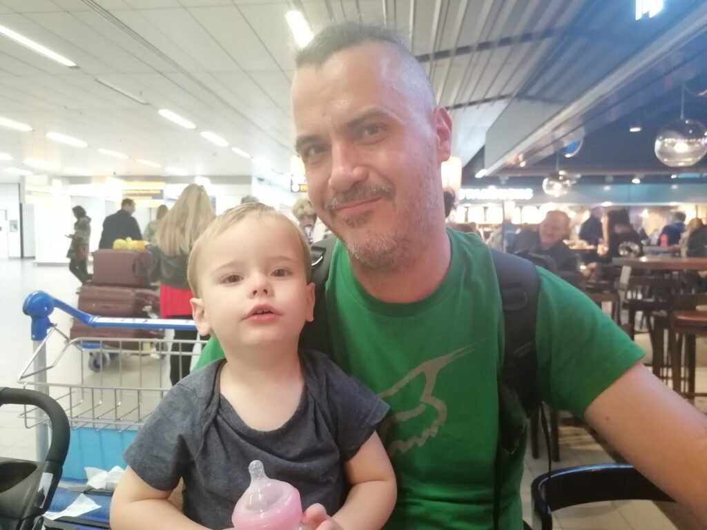 A smiling man wearing a green shirt and a backpack with a toddler on his lap at a restaurant.