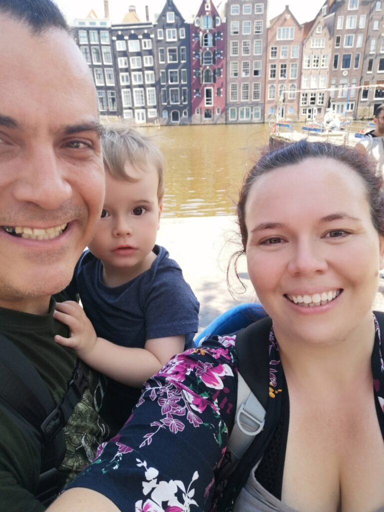 A smiling man and woman holding a toddler while standing in front of a canal in Amsterdam.