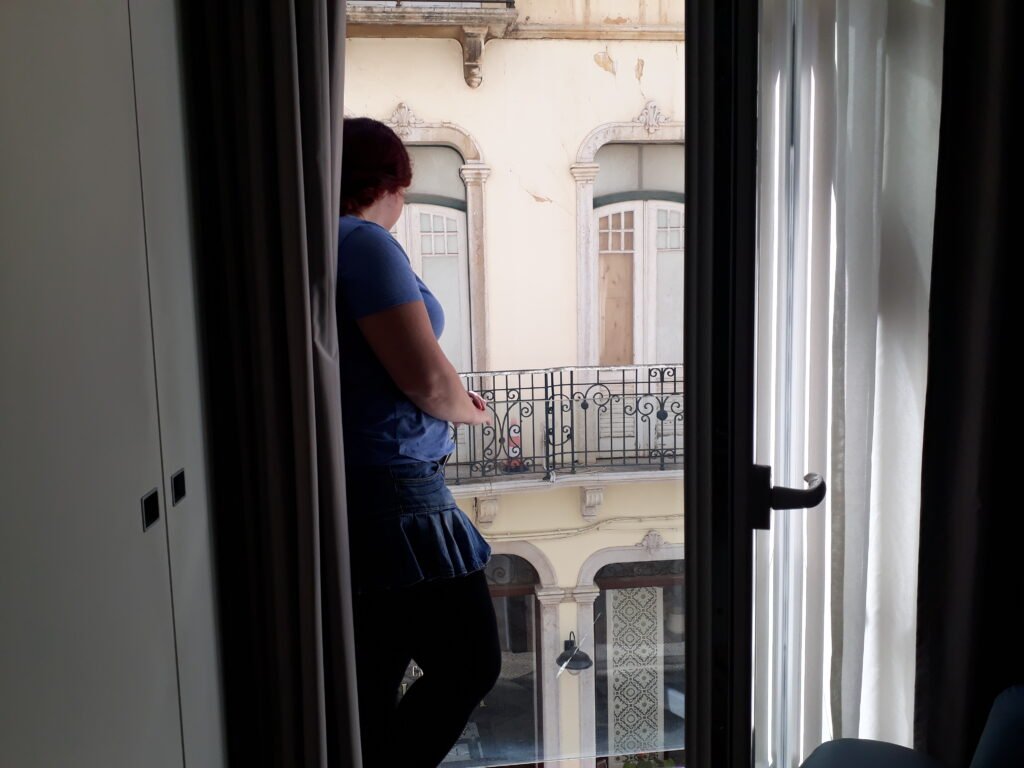 A woman in a skirt and blue shirt standing by a balcony window in a hotel room looking out at the streets of Faro, Portugal