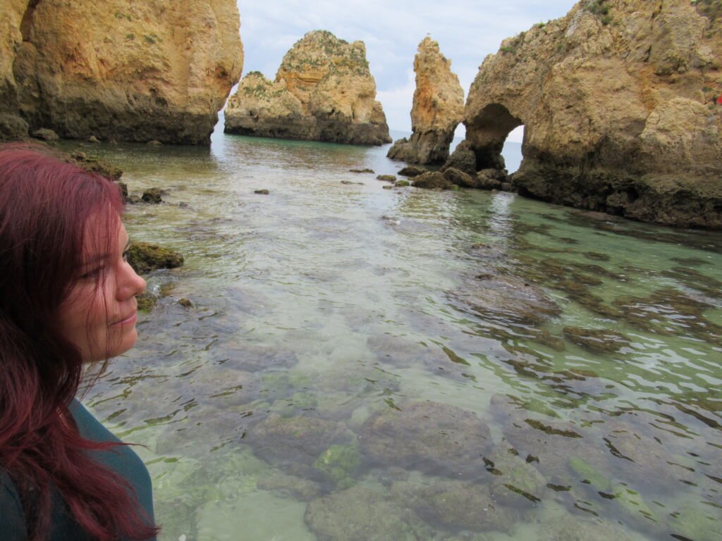 A woman with red hair and a blue shirt standing near the water looking out at the rock cliffs at Lagos's Ponta da Piedade.