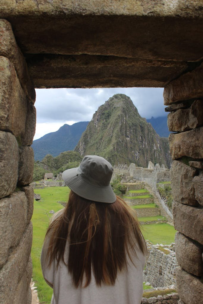 Person with long hair and hat gazing at Machu Picchu through stone window.