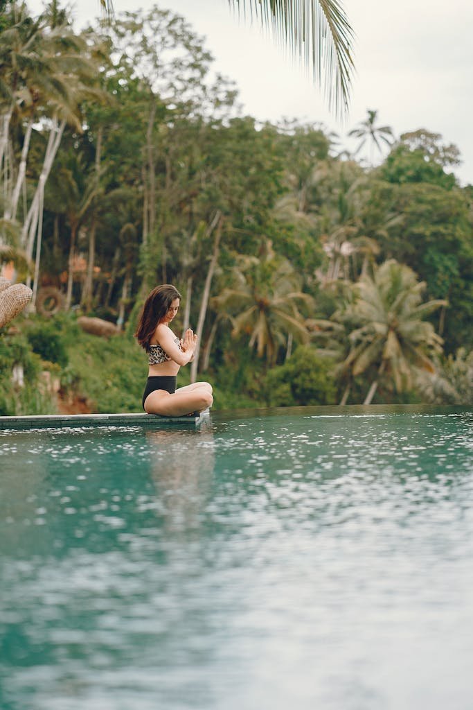 Side view full length of slim unrecognizable brunette in swimwear sitting in lotus pose and meditating while spending time near peaceful pond in tropical country