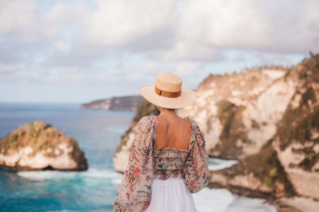 Woman with floral blouse and hat enjoying scenic ocean view from cliff edge.