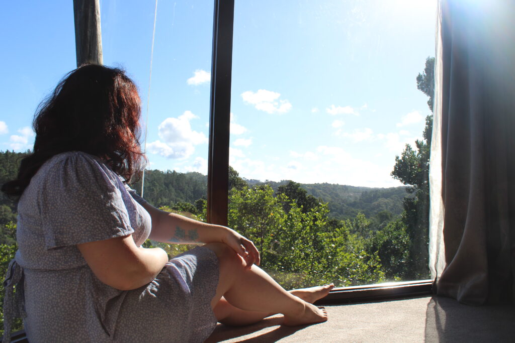 A woman in a blue dress sitting on the carpet looking out a floor to ceiling window at a forest below.