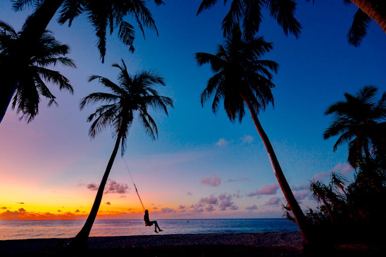A picturesque tropical sunset with a silhouetted person swinging between palm trees on the beach.