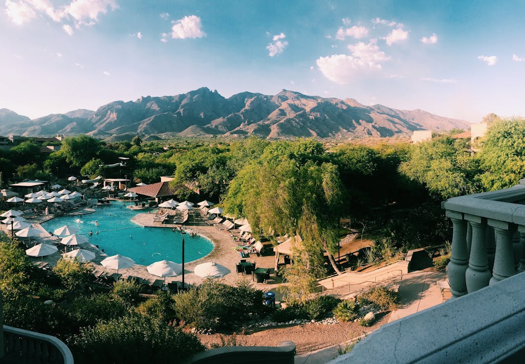 Aerial view of a resort pool surrounded by trees, with mountains in the background under a sunny sky.
