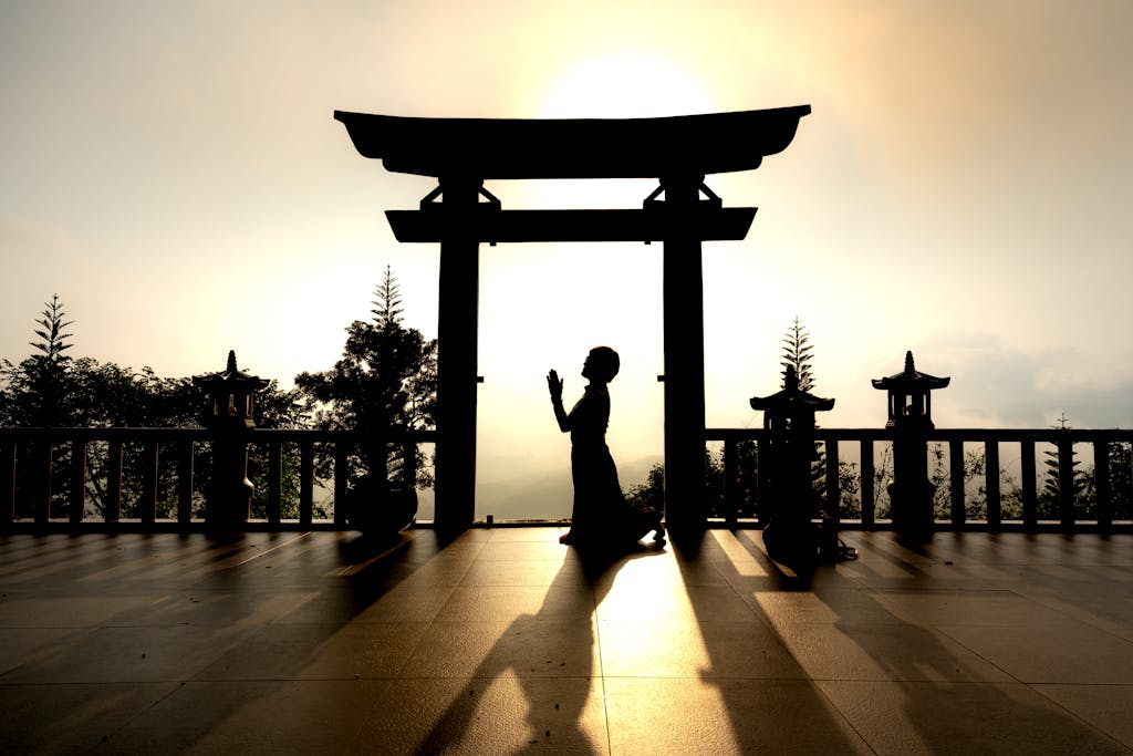 Silhouette of a person kneeling in prayer under a Torii gate at sunrise, symbolizing peace and spirituality.